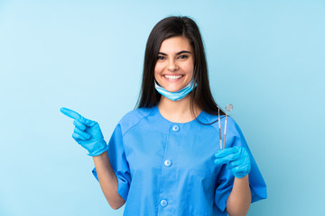 Young woman dentist holding tools over isolated blue background pointing finger to the side