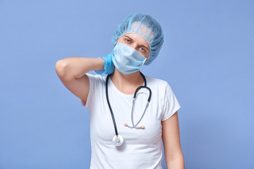 Picture of exhausted hard working young doctor touching her neck, feeling pain, wearing medical hat, gloves and mask, having tough working day, being tired. People and working process concept.