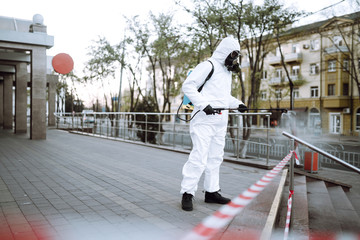 Man in protective suit  and mask sprays disinfector onto the railing in the empty public place at dawn in the city of quarantine. Covid -19. Cleaning concept.