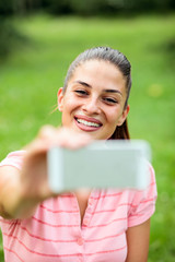 Smiling happy young Caucasian woman resting after a hard workout in the park, taking a selfie with smart phone