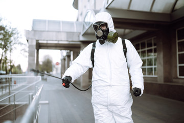 Man in protective suit  and mask sprays disinfector onto the railing in the empty public place at dawn in the city of quarantine. Covid -19. Cleaning concept.