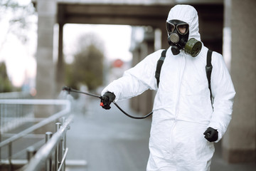 Man in protective suit  and mask sprays disinfector onto the railing in the empty public place at dawn in the city of quarantine. Covid -19. Cleaning concept.