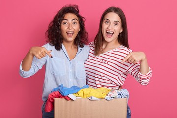 Closeup portrait of two excited girls pointing at carton box with clothes for secondary using,...