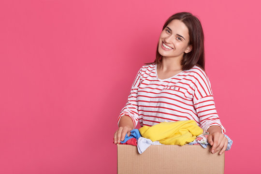 Image of brunette lady with pleasant appearance, standing with box in hands against pink wall, doing charity work, poses with clothes for poor and homeless people. Copy space for advertisement.