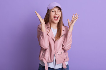 Obraz na płótnie Canvas Studio shot of screaming woman posing with raised hands and widely opened mouth, wearing stylish leather jacket,casual shirt and cap, posing isolated over lilac studio background. People emotions.