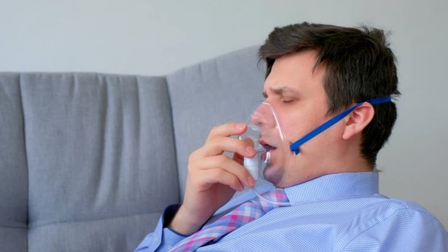 Portrait of sick man in oxygen mask due to difficulty in breathing in coronavirus pandemic sitting on couch at home, side view. Making deep breathes. Breathing problem. Worldwide COVID-19 epidemic.