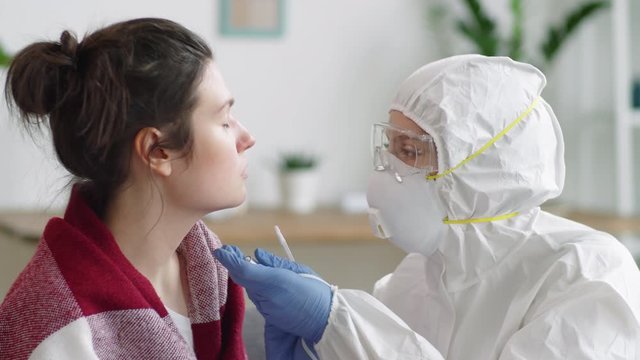 Female doctor in protective suit, gloves, glasses and face mask collecting nasal swab from young woman and talking to her while testing her for virus at home