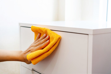 Woman hand cleaning white cabinet with orange color microfiber cloth in bedroom at home. Concept of disinfecting surfaces from bacteria or viruses. Close up