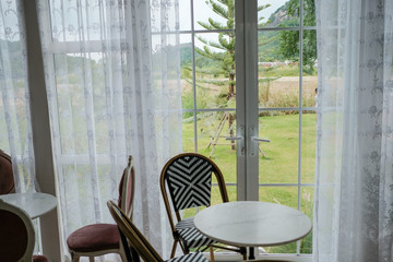 Wood vintage tables and chairs near clear glass doors with field of trees and lake background.