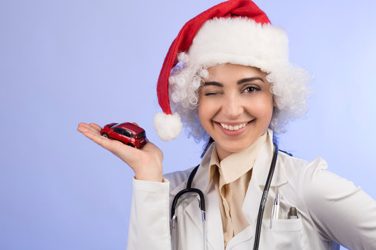 dottoressa in camice bianco , Stetoscopio intorno collo e con il cappello da babbo natale, mostra una machinetta giocattolo rossa,  isolata su sfondo celeste