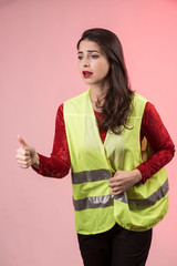 Ragazza mora che indossa una maglia rossa e un gilet giallo ad alta visibilità chiede l'autostop con il pollice ,isolata su sfondo rosa