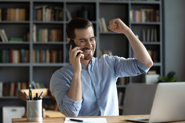 Smiling satisfied businessman wearing glasses making phone call, hearing good news, job promotion celebrating success, showing yes gesture, sitting at work desk with laptop and using smartphone