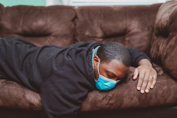 african american man laying down on couch with black sweat shirt wearing a blue surgical mask appearing sick and sleepy