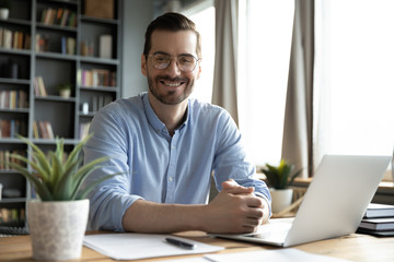 Head shot portrait smiling businessman wearing glasses sitting at work desk with laptop, looking at...