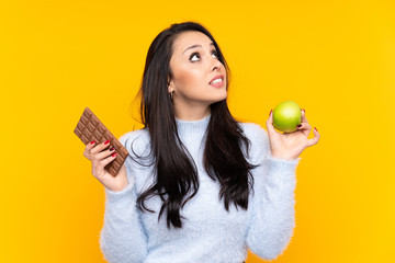 Young Colombian girl over isolated yellow background having doubts while taking a chocolate tablet...