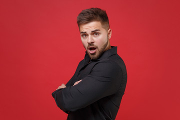 Side view of shocked young bearded guy 20s in classic black shirt posing isolated on red wall background studio. People lifestyle concept. Mock up copy space. Holding hands crossed, looking camera.