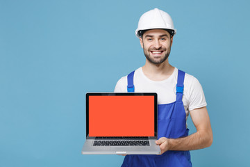 Smiling man in coveralls protective helmet hardhat hold laptop computer with blank empty screen isolated on blue background. Instruments accessories for renovation apartment room. Repair home concept.