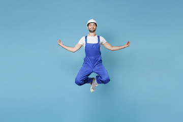 Obraz premium Smiling young man in coveralls hardhat isolated on blue background. Instruments accessories for renovation apartment room. Repair home concept. Jumping hold hands in yoga gesture, relaxing meditating.