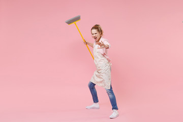 Irritated woman housewife in casual clothes apron doing housework isolated on pink background. Housekeeping concept. Mock up copy space. Sweeping with broom screaming showing stop gesture with palm.