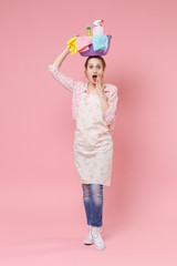 Shocked young woman housewife in apron doing housework isolated on pink wall background studio. Housekeeping concept. Holding basin with detergent bottles washing cleansers on head, put hand on cheek.