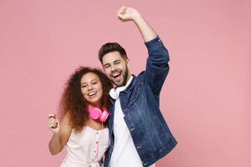 Laughing young couple two friends european guy african american girl in casual clothes with headphones posing isolated on pastel pink background studio. People lifestyle concept. Rising hands dancing.