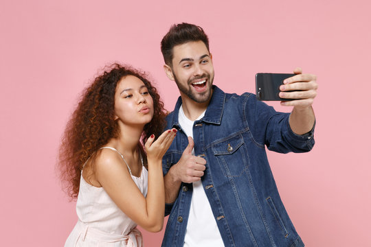 Funny young couple friends european guy african american girl isolated on pastel pink background. People lifestyle concept. Doing selfie shot on mobile phone, blowing send air kiss, showing thumb up.