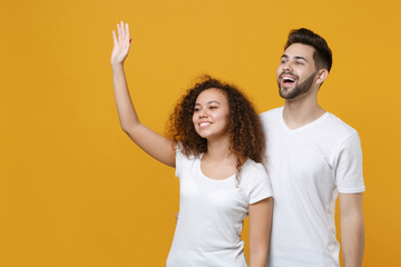 Cheerful couple friends european guy african american girl in white t-shirts isolated on yellow background. People lifestyle concept. Mock up copy space. Waving greeting with hand as notices someone.