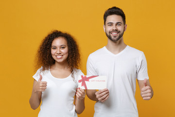 Smiling young couple two friends european guy african american girl in white t-shirts posing isolated on yellow background studio. People lifestyle concept. Hold gift certificate, showing thumbs up.