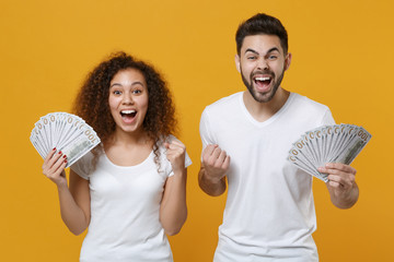 Excited couple friends european guy african american girl in white t-shirts isolated on yellow background. People lifestyle concept. Hold fan of cash money in dollar banknotes, doing winner gesture.