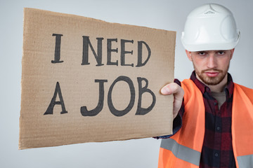 A construction worker holds up a cardboard sign that says I need a job. Unemployment. Bankruptcy of...