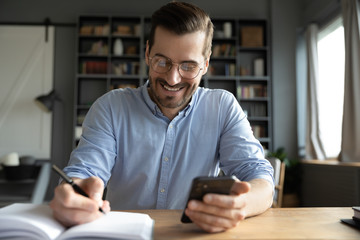 Smiling businessman wearing glasses using phone, writing important information in notebook, sitting at desk, happy man holding smartphone, making notes in personal daily planner, planning work day