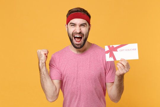 Screaming bearded fitness sporty guy sportsman in headband t-shirt in home gym isolated on yellow background. Workout sport motivation lifestyle concept. Hold gift certificate, doing winner gesture.