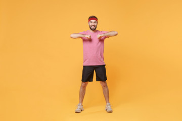Fototapeta na wymiar Cheerful young bearded fitness sporty guy sportsman in headband t-shirt in home gym isolated on yellow background. Workout sport motivation concept. Doing warm up spreading hands with clenching fists.