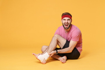 Ingured screaming young bearded fitness sporty guy sportsman in headband t-shirt in home gym isolated on yellow background. Workout sport motivation concept. Sit touch ankle foot with elastic bandage.