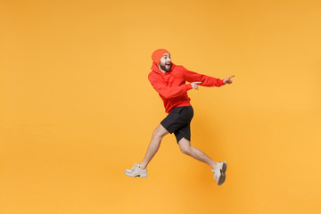 Fototapeta na wymiar Excited young bearded fitness sporty guy 20s sportsman in hat hoodie shorts in home gym isolated on yellow background. Workout sport motivation lifestyle concept. Jumping pointing index fingers aside.