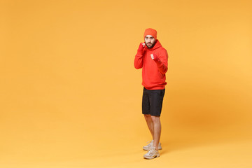 Young bearded fitness sporty guy boxer sportsman in hat, hoodie, shorts spend weekend in home gym isolated on yellow background. Workout sport motivation concept. Boxing with sports bandages on hands.