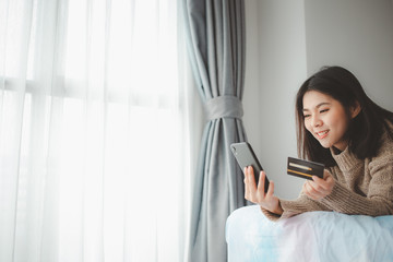 Happy Asian women lying on the bed and using smartphone for online shopping while holding credit card in hand