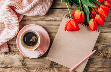 Pink cup of coffee, knitted plaid, clean notebook and real tulip flowers on a rustic wooden background. Female workplace with cozy breakfast
