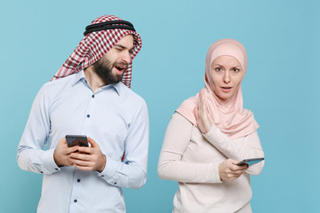 Shocked concerned couple friends arabian muslim man wonam in keffiyeh kafiya ring igal agal hijab clothes isolated on blue background. People religious concept. Using mobile phone, typing sms message.