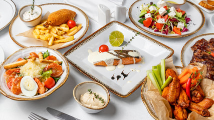 Assorted dishes on table. Salmon steak on the grill, chicken wings, caesar with shrimps, kiev cutlet with fries, greek salad