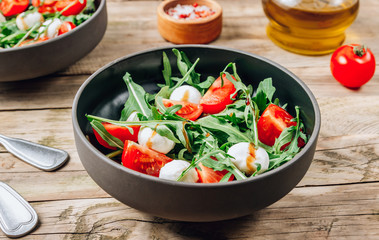 Healthy arugula salad with mozzarella cheese and cherry tomatoes in black bowls on wooden rustic background.