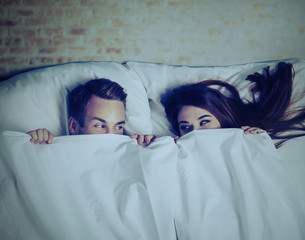 Young adult playful couple peeping from bedsheet on the bed at bedroom. Looking at each other....