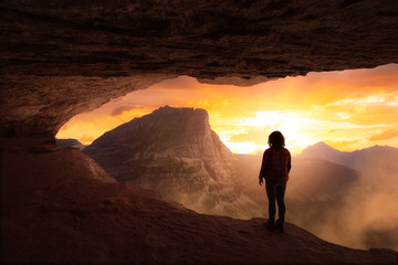 Adventurous Girl Looking out at a dramatic American Mountain Landscape from a rocky cave during a colorful sunset. Fantasy Adventure Composite.