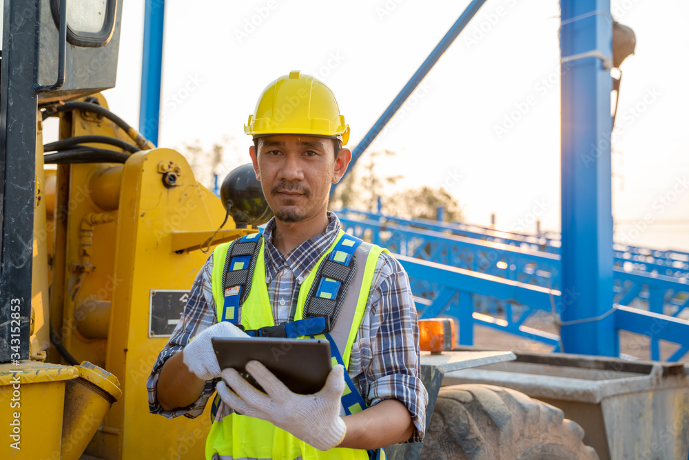 Poster engineering wearing a yellow safety helmet standing in front of the backhoe and are using tablet to 