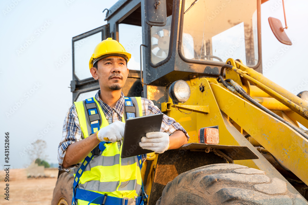 Poster engineering wearing a yellow safety helmet standing in front of the backhoe and are using tablet to 