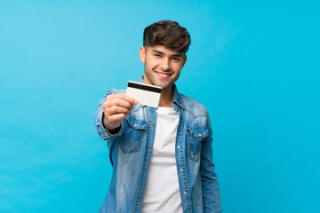 Young handsome man over isolated blue background holding a credit card - obrazy, fototapety, plakaty
