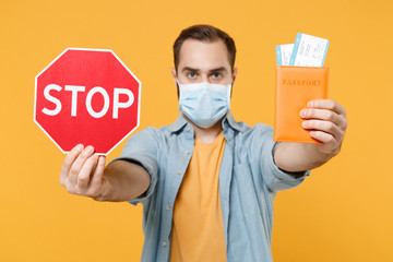 Young man in sterile face mask posing isolated on yellow background in studio. Epidemic pandemic coronavirus 2019-ncov sars covid-19 flu virus concept. Hold passport tickets, red sign with Stop title.