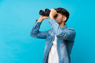 Young handsome man over isolated blue background with black binoculars