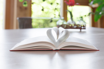 Zwei Blätter eines aufgeschlagenen Buches bilden eine Herzform. Das Buch liegt auf einem hellen Tisch und durch Fenster sieht man grüne Natur.