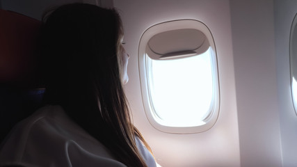young lady with long brown loose hair wearing grey t-shirt looks out bright airliner porthole in passenger cabin closeup